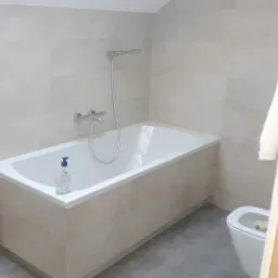 Wykończona łazienka na poddaszu z wanną obudowaną płytkami, szarą podłogą, toaletą i oknem dachowym.