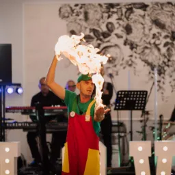 Mężczyzna w kolorowym stroju prezentuje płonący rekwizyt na scenie z zespołem muzycznym w tle, oświetlenie sceniczne i dekoracja kwiatowa na ścianie.