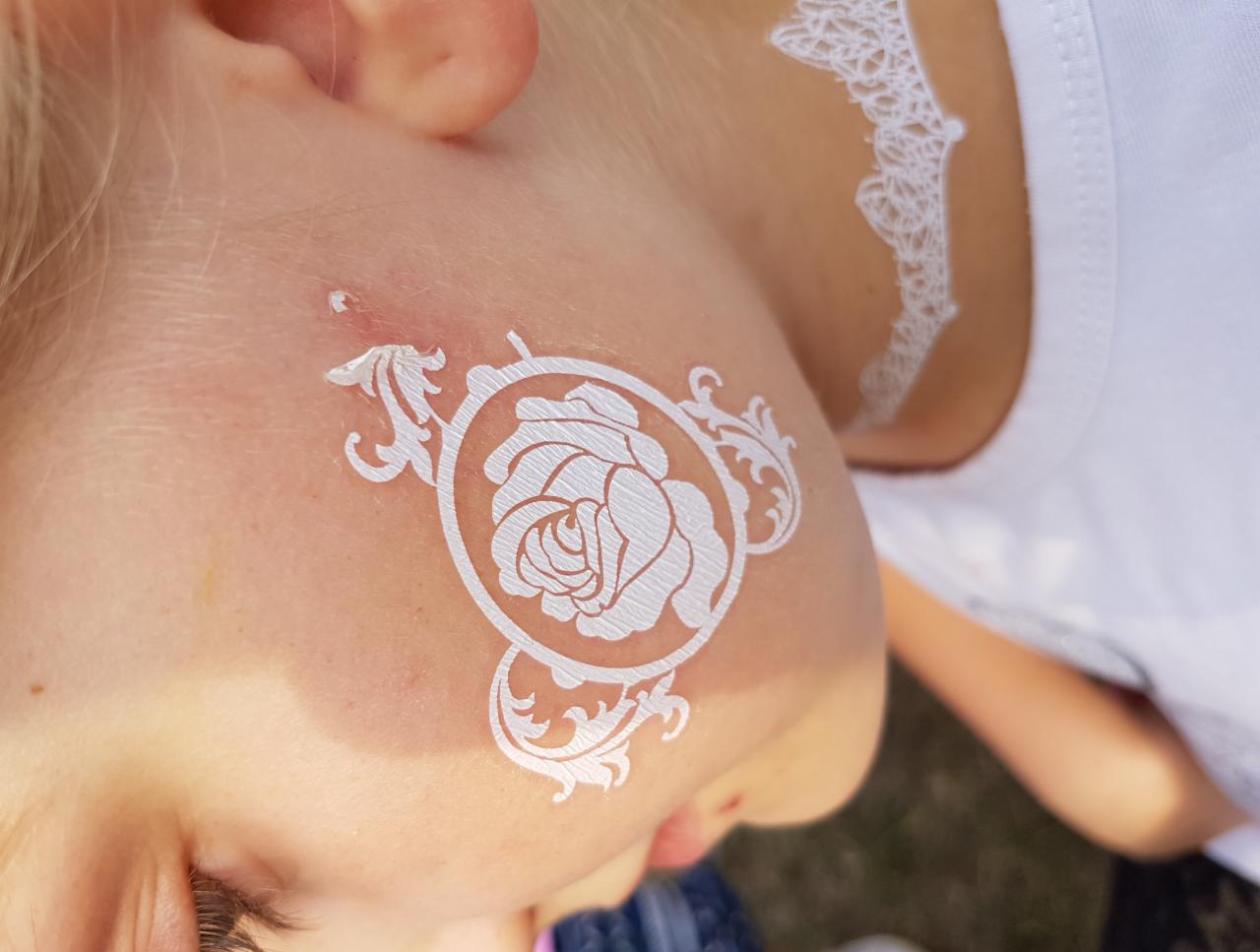 Dziewczynka z białym, ozdobnym tatuażem z motywem róży na policzku i koronkowym wzorem na ramieniu, widok z góry.