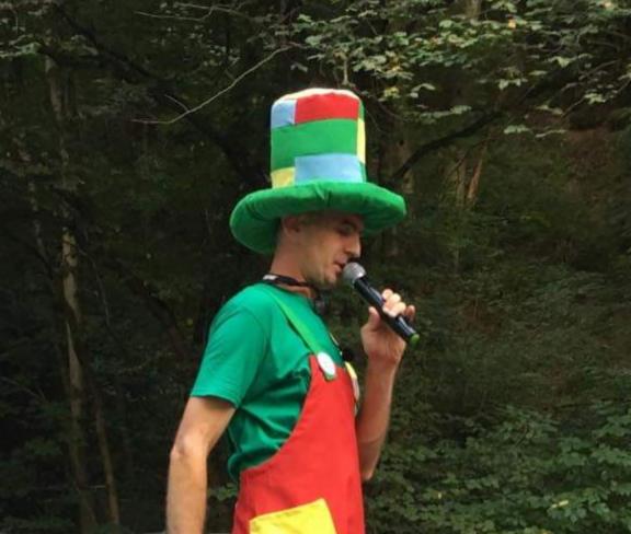 Mężczyzna w kolorowym kostiumie i kapeluszu, trzymający mikrofon, występuje na tle zieleni w Gorlicach.