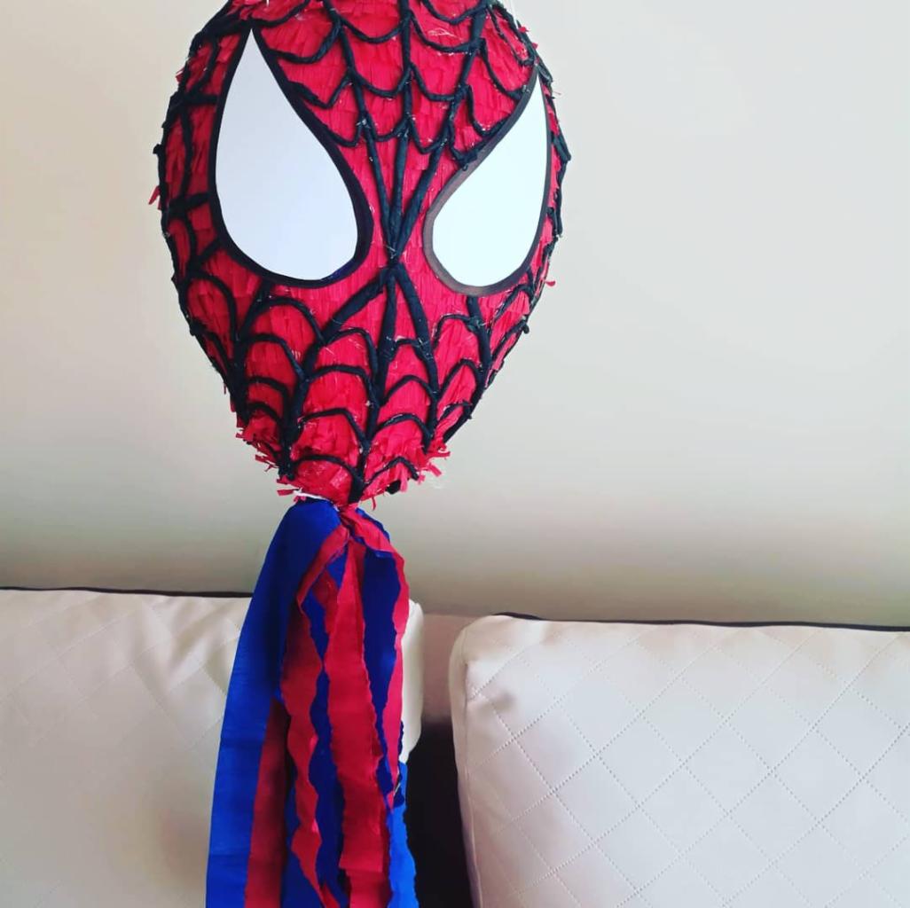 Piñata w kształcie głowy Spider-Mana, wykonana z czerwonej bibuły z czarną siatką i białymi oczami, zwisająca z sufitu, z doczepionymi niebiesko-czerwonymi frędzlami z bibuły.