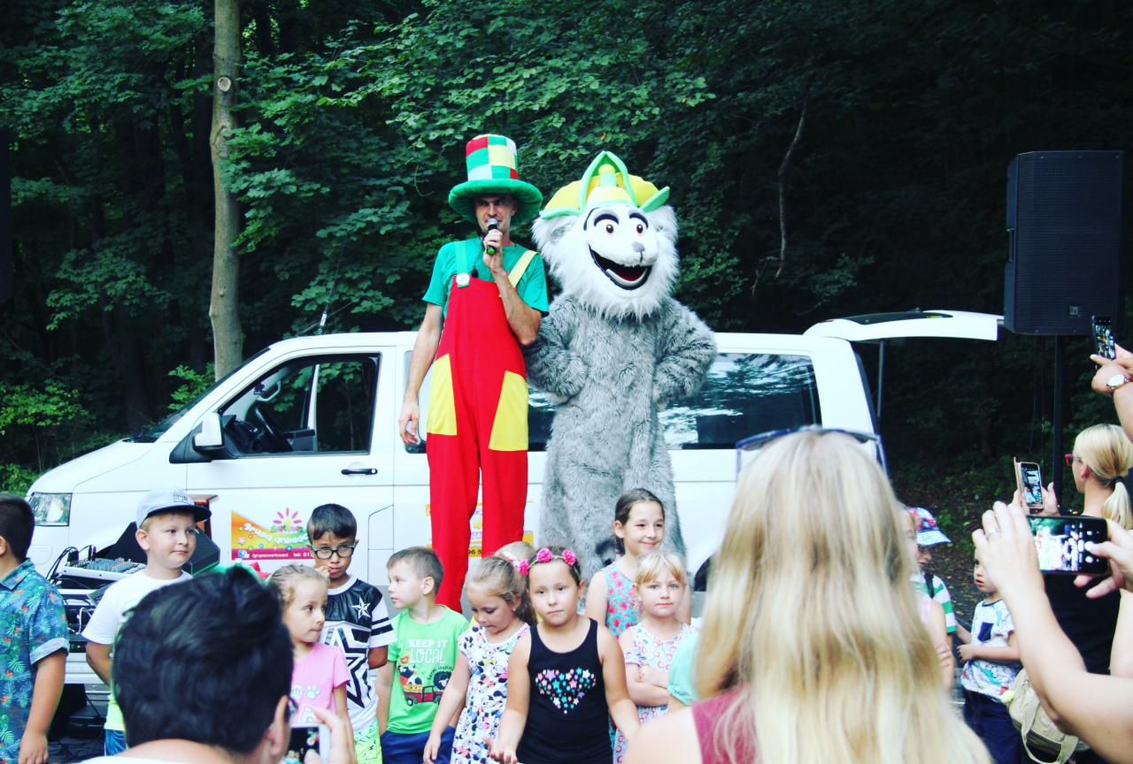 Animator w kolorowym stroju i maskotka w kostiumie zwierzęcia na scenie przed grupą dzieci, z białym vanem i zielonym tłem z drzew