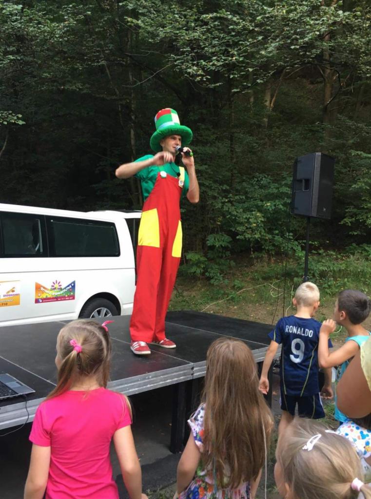 Mężczyzna w kostiumie scenicznym z zielonym kapeluszem i czerwono-żółtym kombinezonie, śpiewa do mikrofonu na scenie podczas występu dla dzieci na tle zieleni.