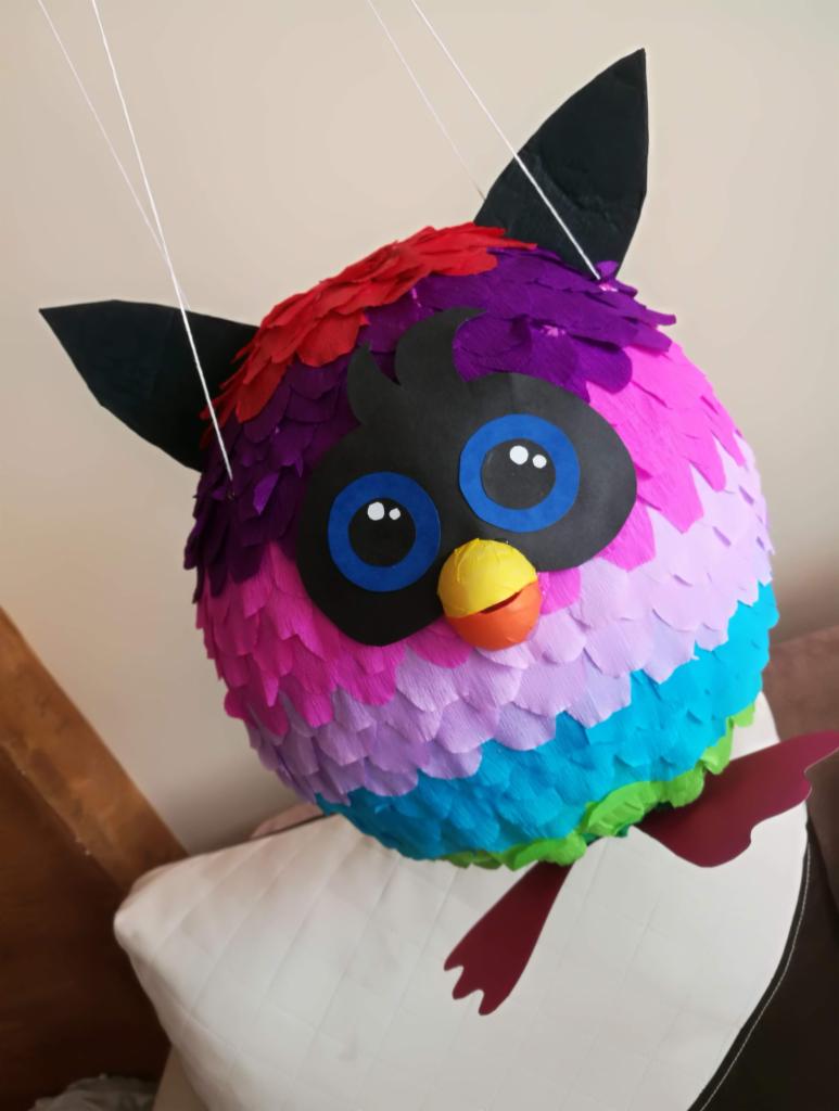 Ręcznie robiona pinata w kształcie sowy Furby z papieru krepowanego w odcieniach czerwieni, fioletu, różu, błękitu i zieleni, zawieszona na białych sznurkach, z czarnymi uszami i oczami...