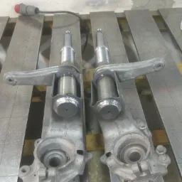 Dwa metalowe elementy zawieszenia samochodowego po procesie łączenia, widoczne ślady spawania na łączeniu cylindrycznych elementów, ułożone na metalowym stole warsztatowym.