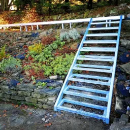 Niebieska metalowa konstrukcja schodów na zboczu skalnym z roślinnością, częściowo pomalowana, widoczne ślady użytkowania, w tle barierka.