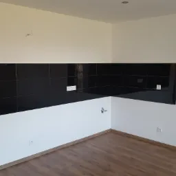 Fragment kuchni po remoncie: czarne płytki na ścianie, białe ściany, drewnopodobna podłoga, widoczne gniazdka elektryczne i instalacja wodna w rogu.