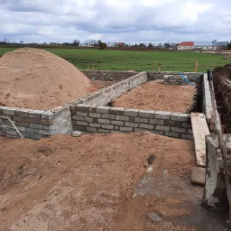 Budowa fundamentów z bloczków betonowych, duża pryzma piasku wewnątrz, wykop pod instalacje widoczny z boku, krajobraz wiejski w tle.