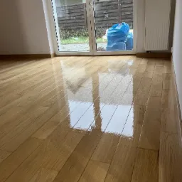 Błyszcząca, świeżo wykończona podłoga z drewnianych desek, odbijająca światło z okna, z widocznymi workami z odpadami budowlanymi za oknem.