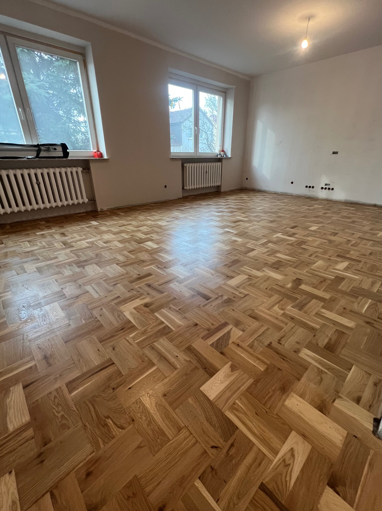 Jasny pokój z nowo ułożonym parkietem w jodełkę, widoczne grzejniki pod oknami i ślady po gniazdkach elektrycznych na ścianie.
