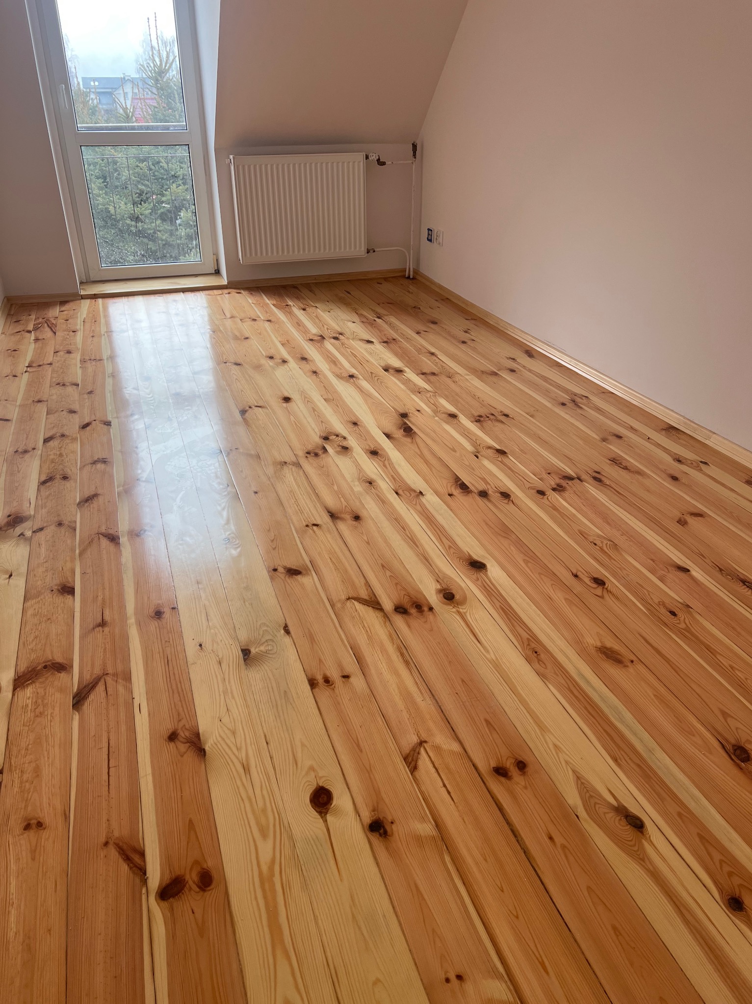 Wypolerowana, jasna podłoga z desek w pokoju na poddaszu, widoczne sęki i naturalny rysunek drewna, okno z widokiem na drzewa na zewnątrz.