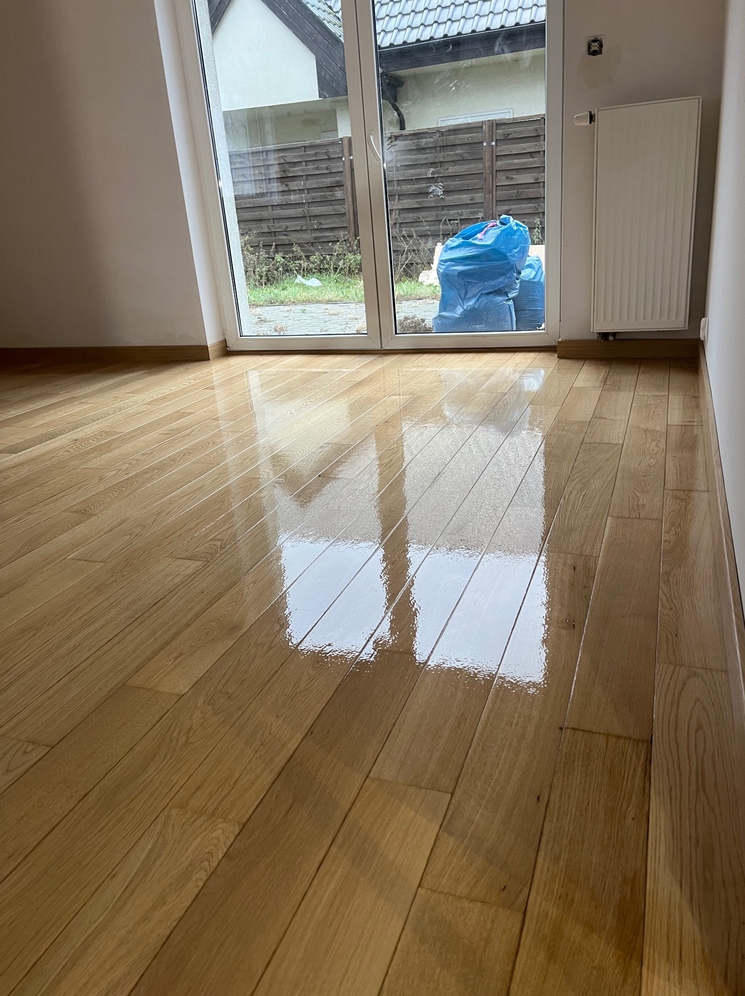 Błyszcząca, świeżo wykończona podłoga z drewnianych desek, odbijająca światło z okna, z widocznymi workami z odpadami budowlanymi za oknem.