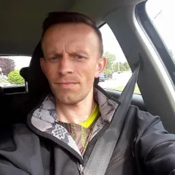 Mężczyzna w samochodzie, zapięty pasem bezpieczeństwa, patrzy wprost. Widoczne drzewa za oknem. Ubrany w czarną kurtkę i kolorową koszulkę.