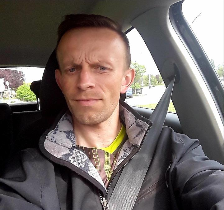 Mężczyzna w samochodzie, zapięty pasem bezpieczeństwa, patrzy wprost. Widoczne drzewa za oknem. Ubrany w czarną kurtkę i kolorową koszulkę.