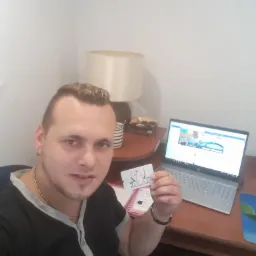 Mężczyzna trzyma wizytówkę z logo firmy, obok laptop z otwartą stroną internetową. Fotografia typu selfie w domowym biurze, z lampą i notatnikiem na biurku.