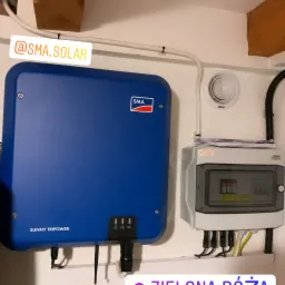 Niebieski falownik SMA Sunny Tripower zamontowany na białej ścianie obok szarej skrzynki elektrycznej Keno z widocznymi kablami, całość sfotografowana z bliska w pomieszczeniu o surowym wykończeniu.