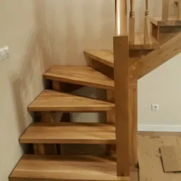 Drewniane schody zabiegowe z jasnego drewna dębowego, z balustradą o toczonych tralkach, zamontowane w narożniku pomieszczenia o jasnych ścianach, z widocznymi elementami zabezpieczającymi podłogę.