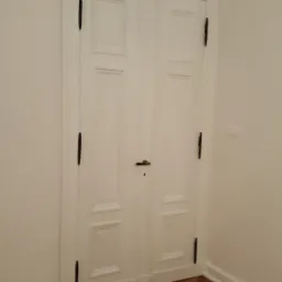 Białe, dwuskrzydłowe drzwi wewnętrzne z czarnymi zawiasami i mosiężną klamką, wkomponowane w białe ściany i parkietową podłogę ułożoną we wzór jodełki.