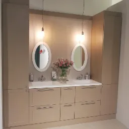 Beżowa łazienka z dwoma owalnymi lustrami, białym blatem i szafkami z szufladami, oświetlona wiszącymi lampami, z wazonem z różowymi kwiatami na blacie.
