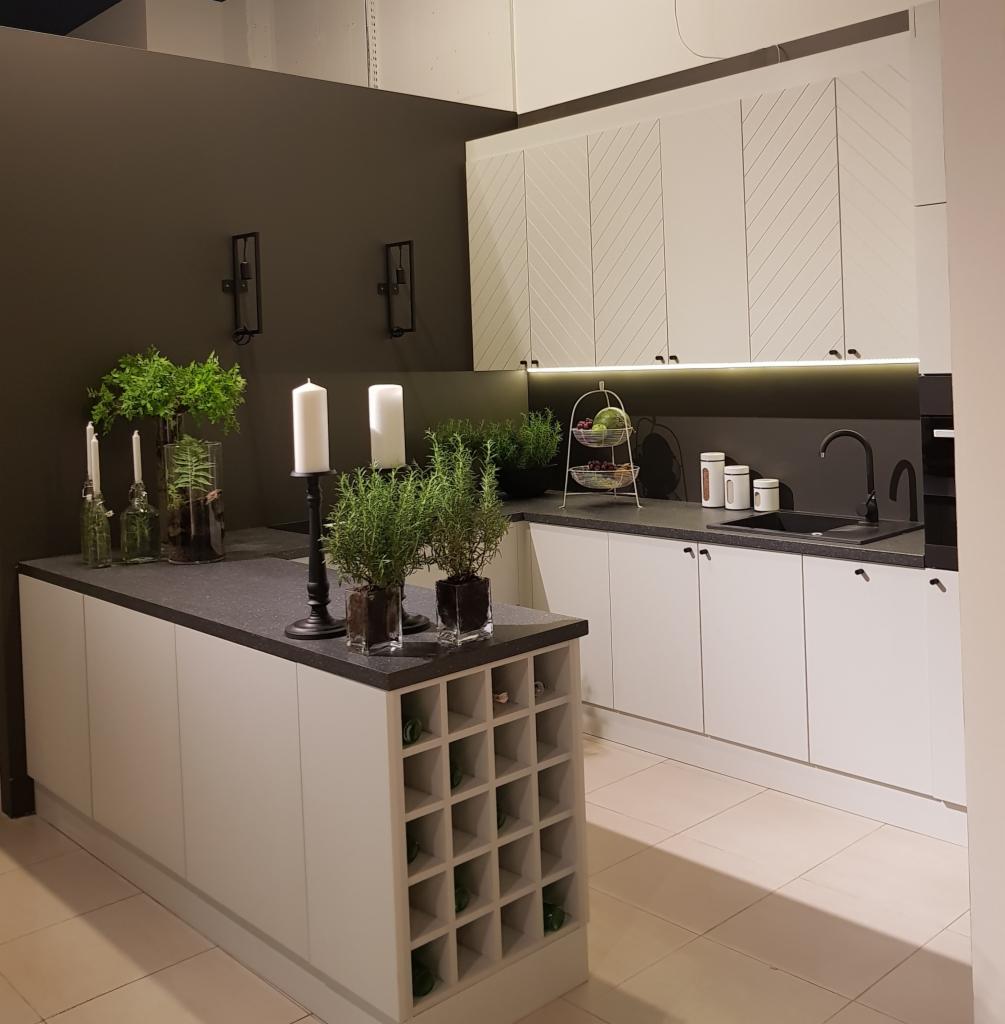Biała kuchnia z wyspą i wbudowanym regałem na wino, czarny blat, białe szafki górne z dekoracyjnym wzorem w jodełkę, nowoczesne oświetlenie podszafkowe, dekoracje roślinne i świece na blacie.