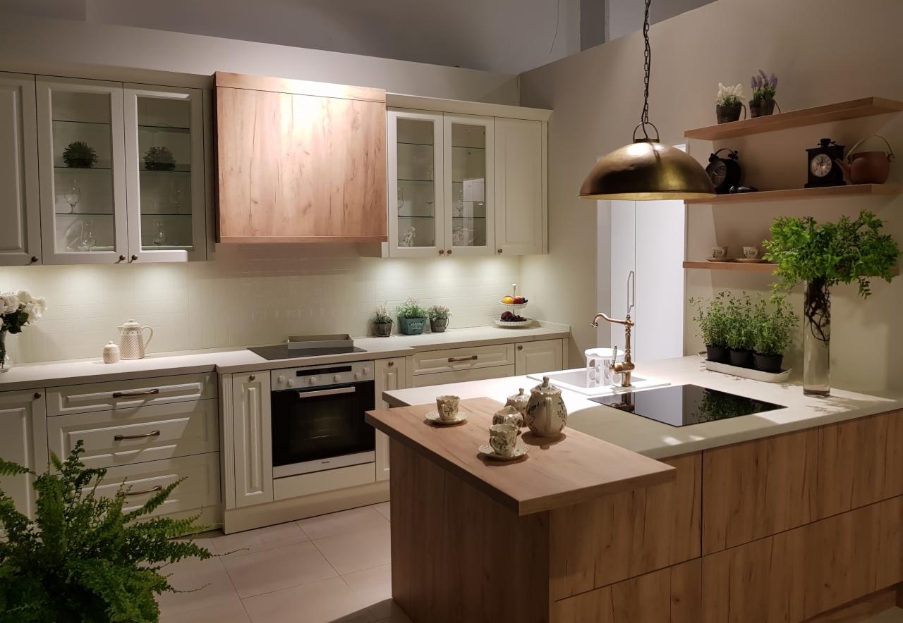 Jasna kuchnia w stylu klasycznym z drewnianymi akcentami, wyspą z blatem i dekoracyjną zastawą do herbaty, oświetlona wiszącą lampą w kolorze starego złota.