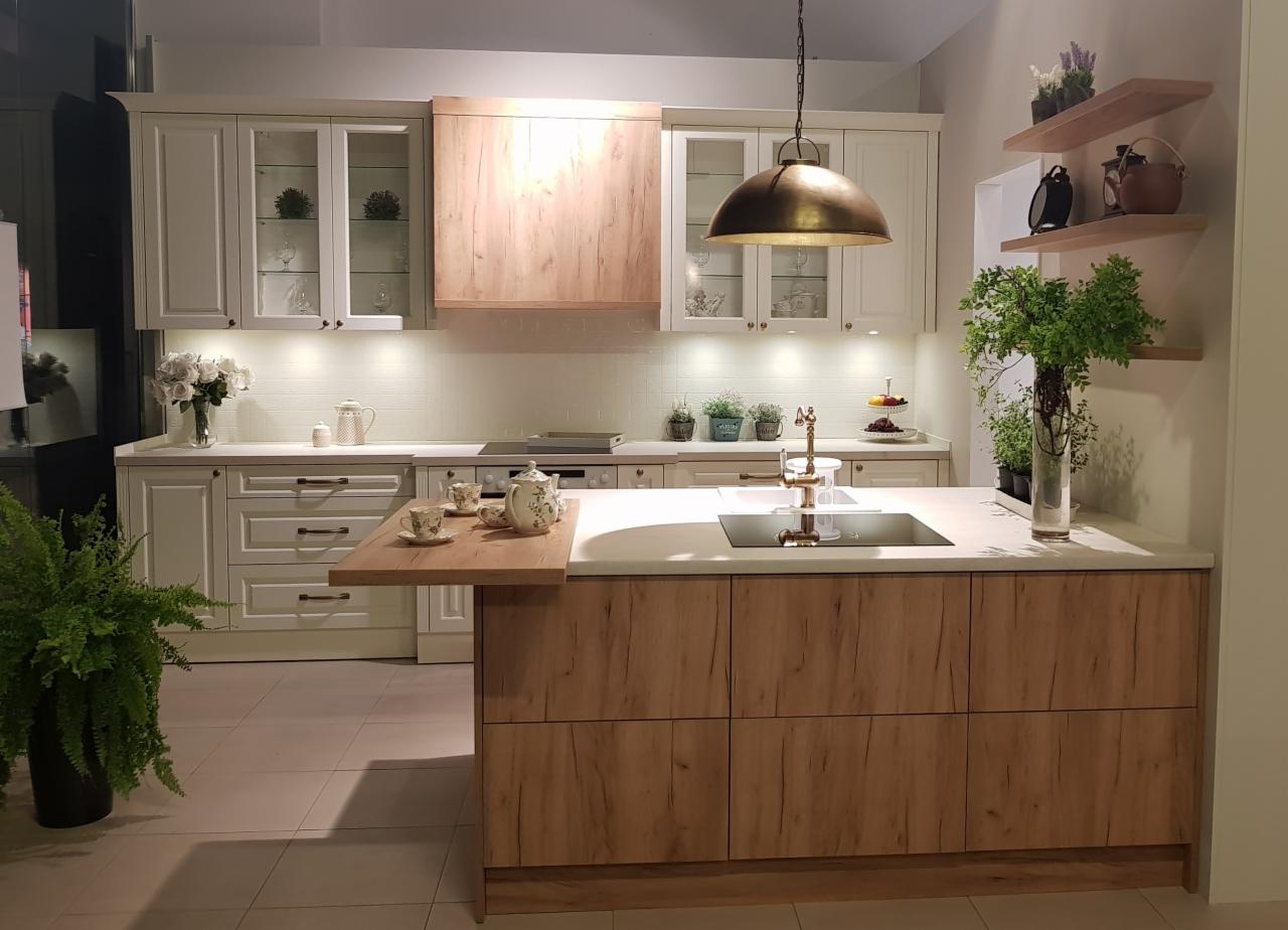 Jasna kuchnia z wyspą, drewnianymi frontami szafek i blatem z konglomeratu, oświetlona wiszącą lampą w stylu retro, z dodatkami w postaci roślin i porcelanowego serwisu do herbaty.