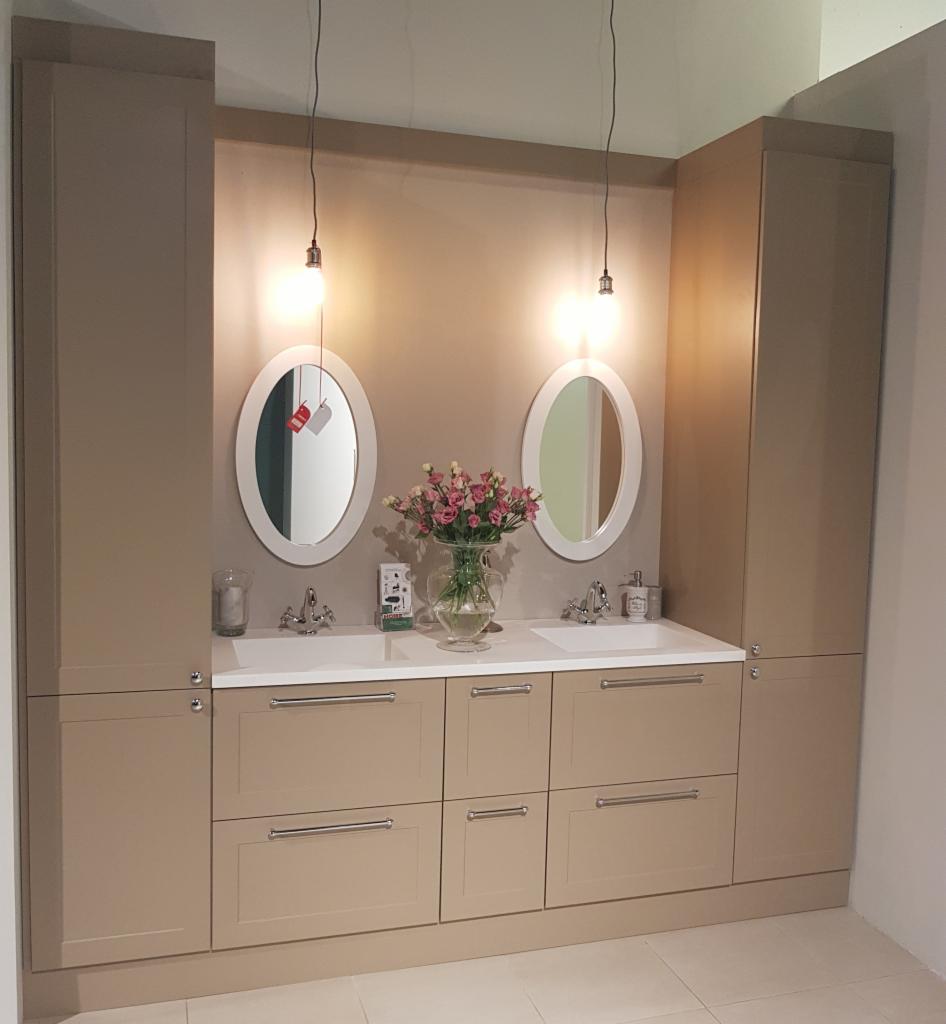 Beżowa łazienka z dwoma owalnymi lustrami, białym blatem i szafkami z szufladami, oświetlona wiszącymi lampami, z wazonem z różowymi kwiatami na blacie.