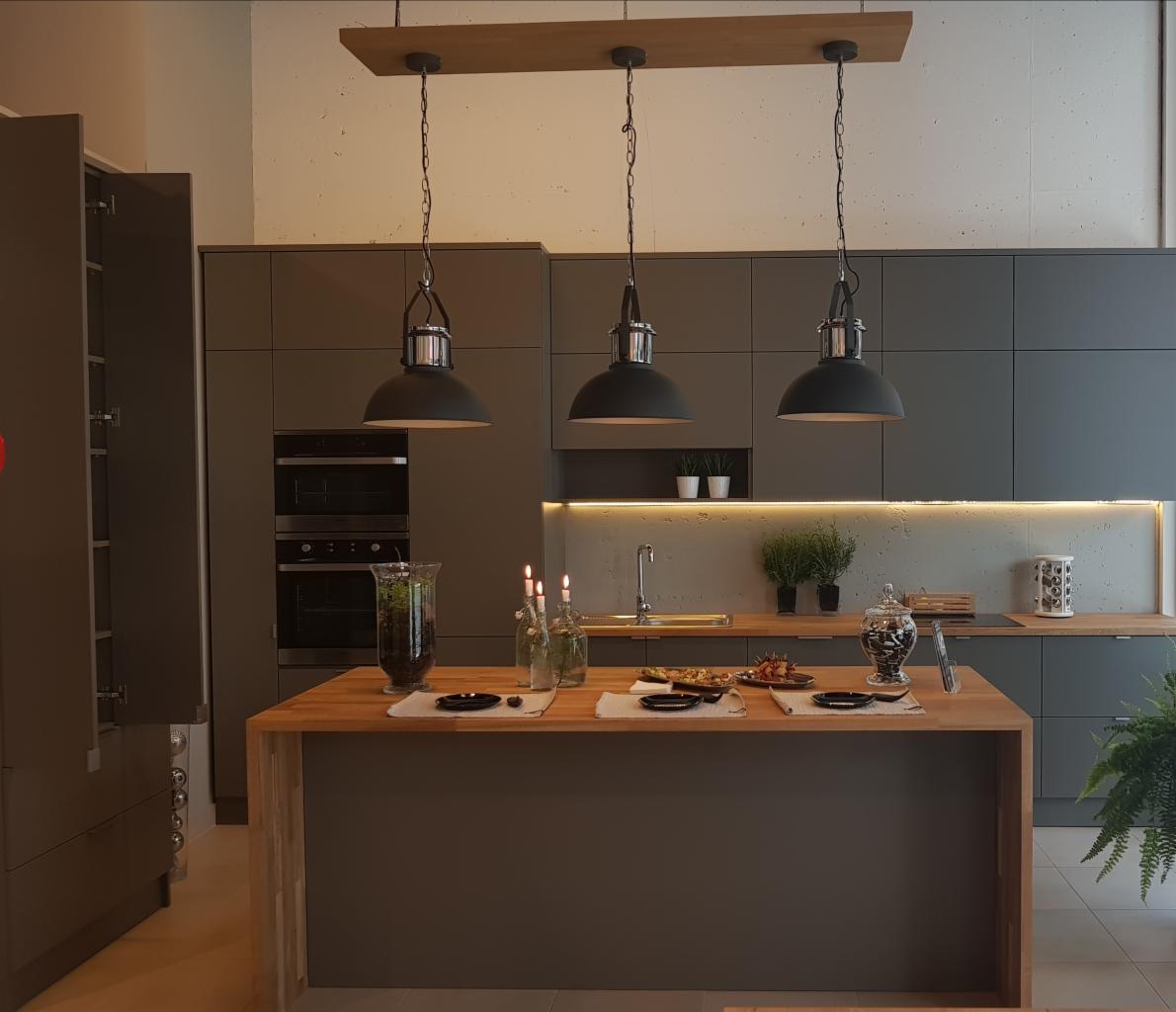 Szara kuchnia z wyspą z drewnianym blatem, oświetlona trzema wiszącymi lampami w stylu industrialnym, dekoracje na blacie, w tle zabudowa kuchenna z piekarnikiem.