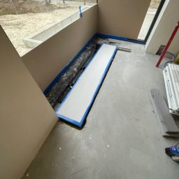 PLOMERO SPÓŁKA Z OGRANICZONĄ ODPOWIEDZIALNOŚCIĄ - Przygotowanie podłoża pod ogrzewanie podłogowe w nowoczesnym wnętrzu z dużym oknem, widoczne rury w izolacji, płyta styropianowa z niebieską taśmą, poziomica, grzejnik i butelka Pepsi.