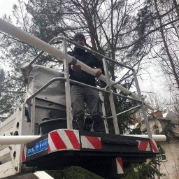 PLOMERO SPÓŁKA Z OGRANICZONĄ ODPOWIEDZIALNOŚCIĄ - Pracownik na podnośniku koszowym w Warszawie instaluje rury, prawdopodobnie wentylacyjne, na tle drzew i budynku.