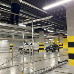 PLOMERO SPÓŁKA Z OGRANICZONĄ ODPOWIEDZIALNOŚCIĄ - Instalator na rusztowaniu montuje rury pod sufitem parkingu podziemnego, widoczne elementy instalacji i narzędzia pomiarowe.