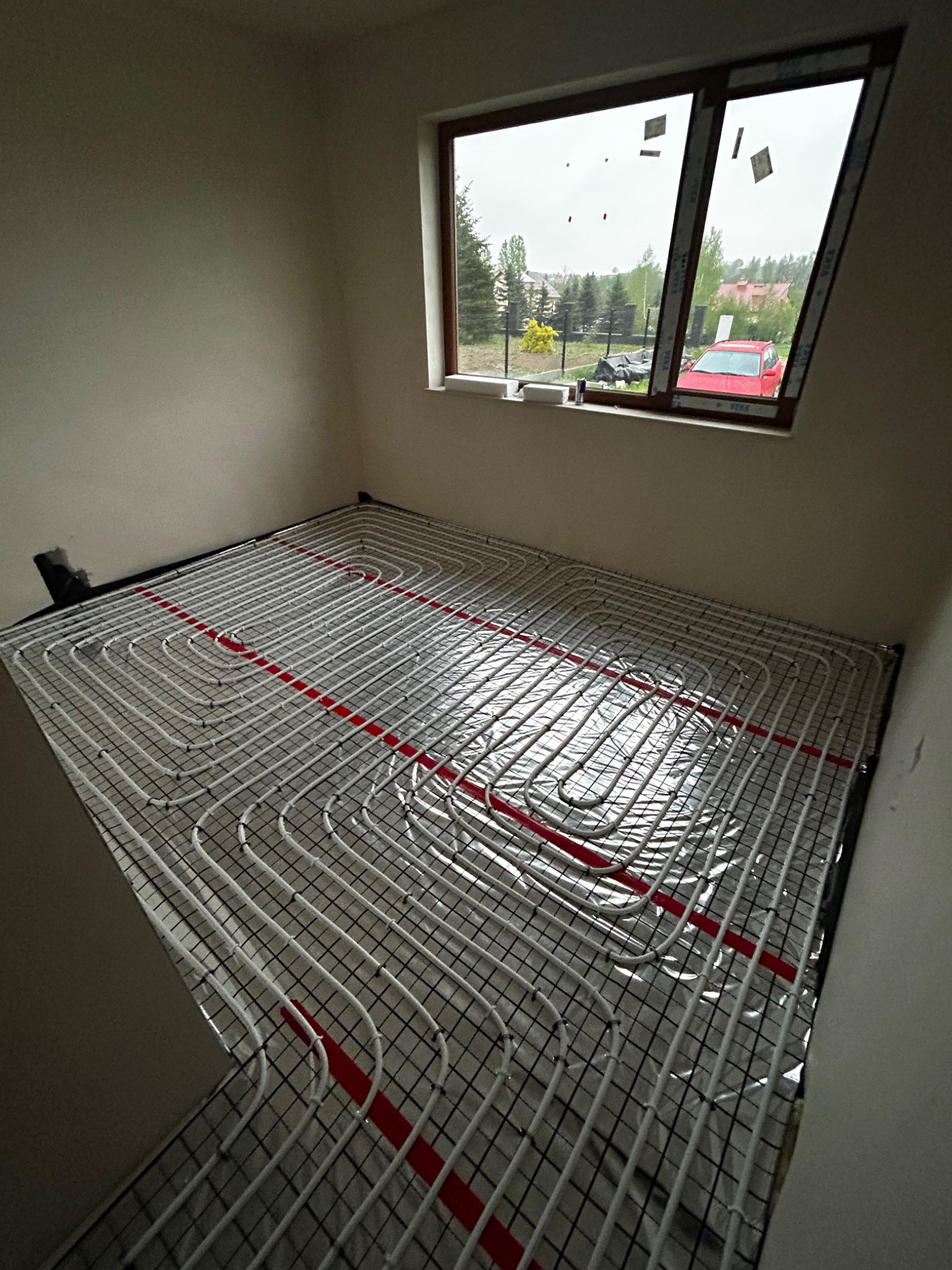 Białe rury ogrzewania podłogowego ułożone na srebrnej folii z czarną siatką, z czerwonymi elementami mocującymi, widoczne w pustym pomieszczeniu z oknem, przez które widać dom i czerwony samochód.