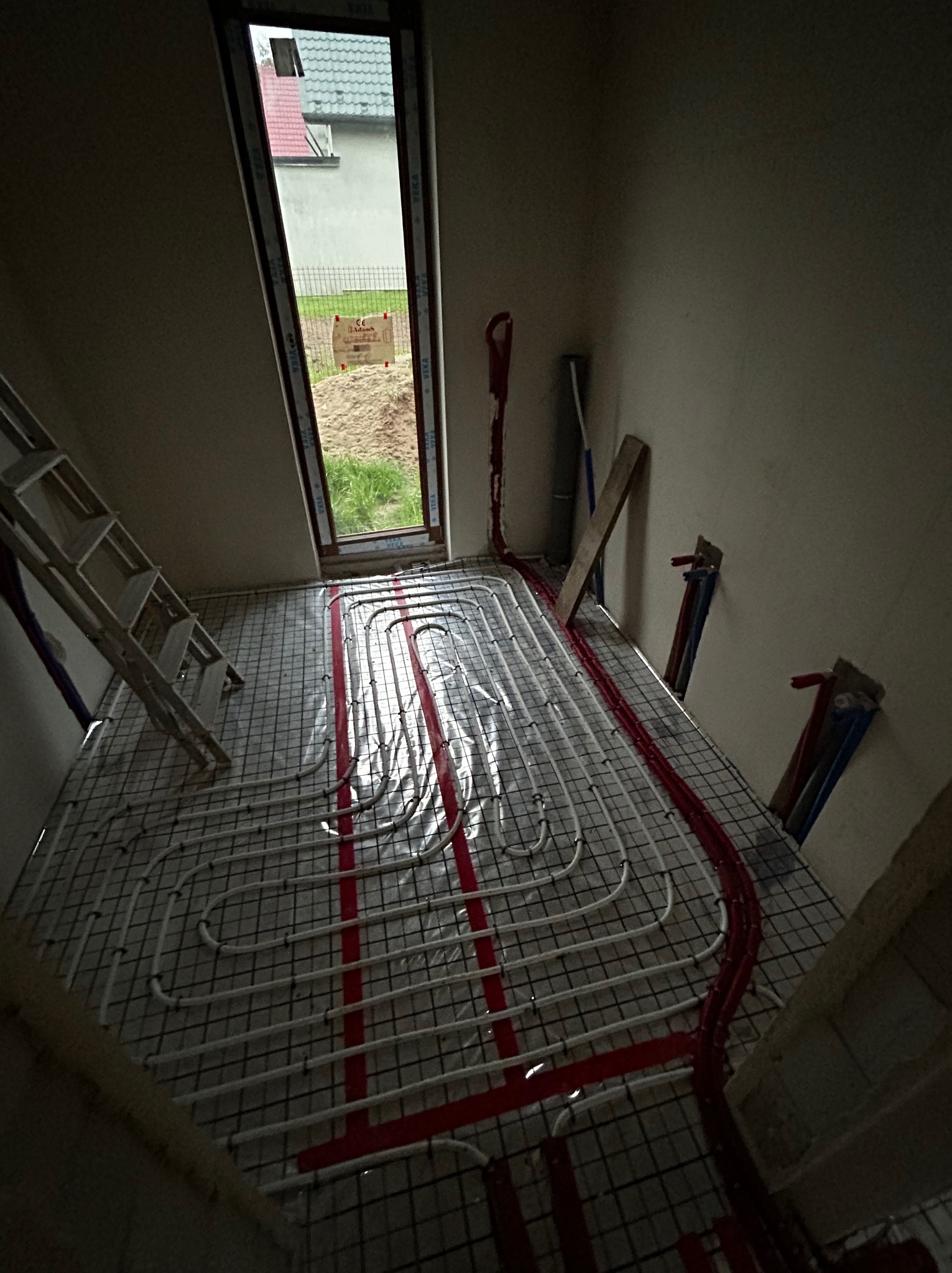 Montaż ogrzewania podłogowego w trakcie budowy w Warszawie: widoczne białe rurki na siatce, czerwone elementy mocujące, drabina oparta o ścianę, okno z widokiem na ogród.