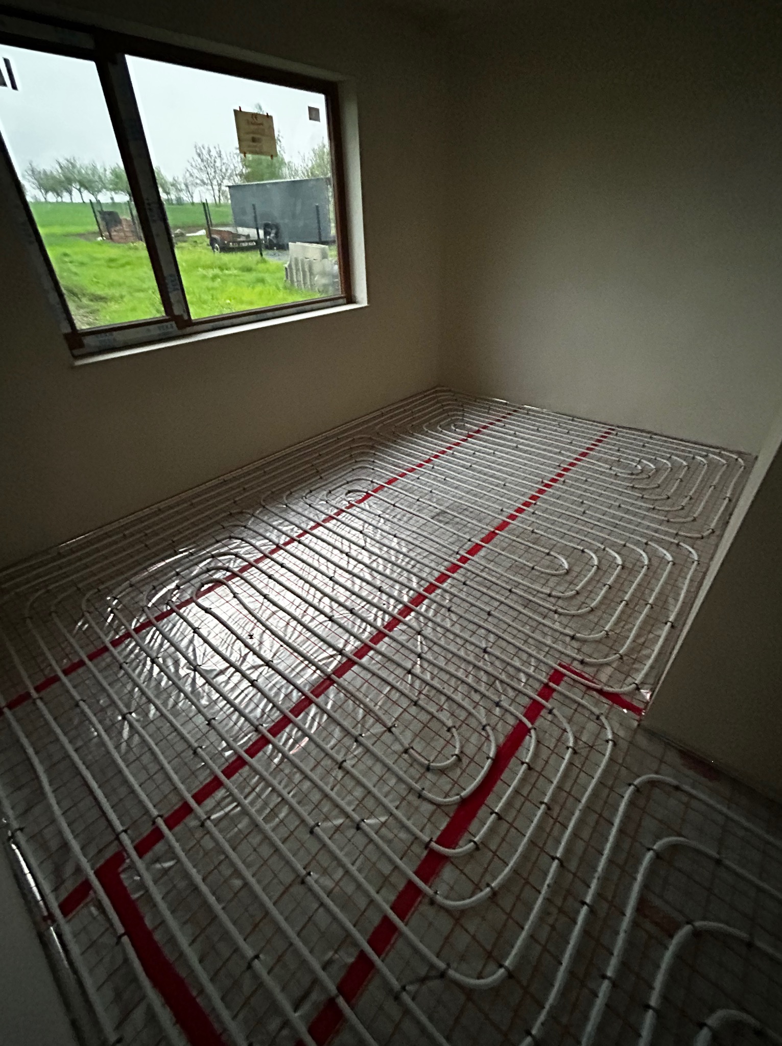 Białe rury ogrzewania podłogowego ułożone na folii z czerwoną siatką, widok z góry, w tle okno z widokiem na pochmurne pole.