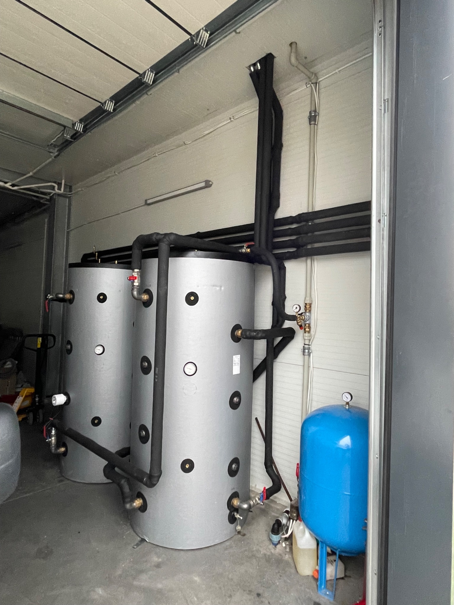 Dwa srebrne zbiorniki buforowe z czarnymi rurami izolowanymi, niebieskie naczynie wzbiorcze, zamontowane w kotłowni z blachy falistej.