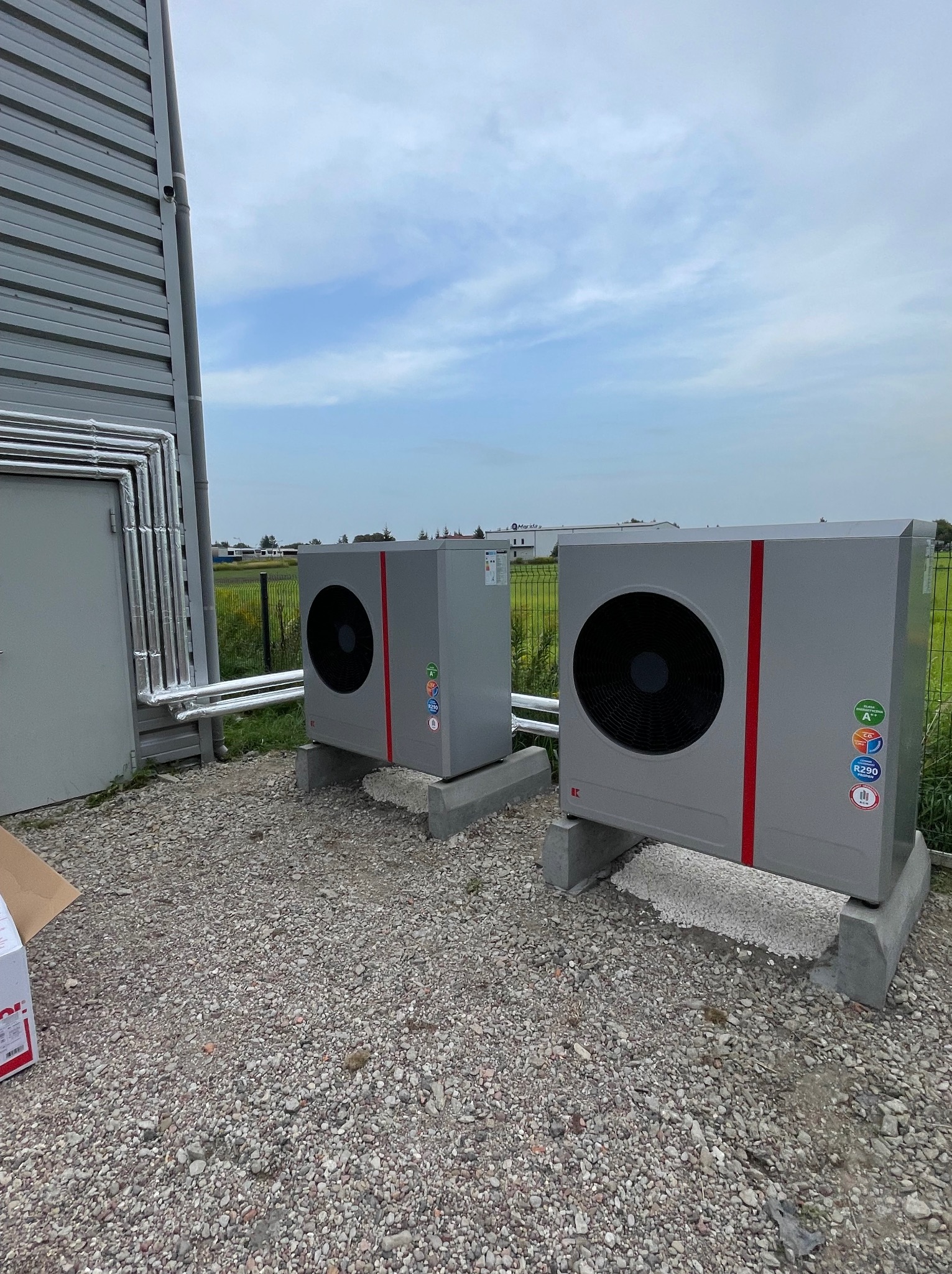 Dwa szare, nowoczesne pompy ciepła z czerwonymi akcentami, ustawione na betonowych podstawach na żwirowym podłożu obok budynku z metalową elewacją i zaizolowanymi rurami.