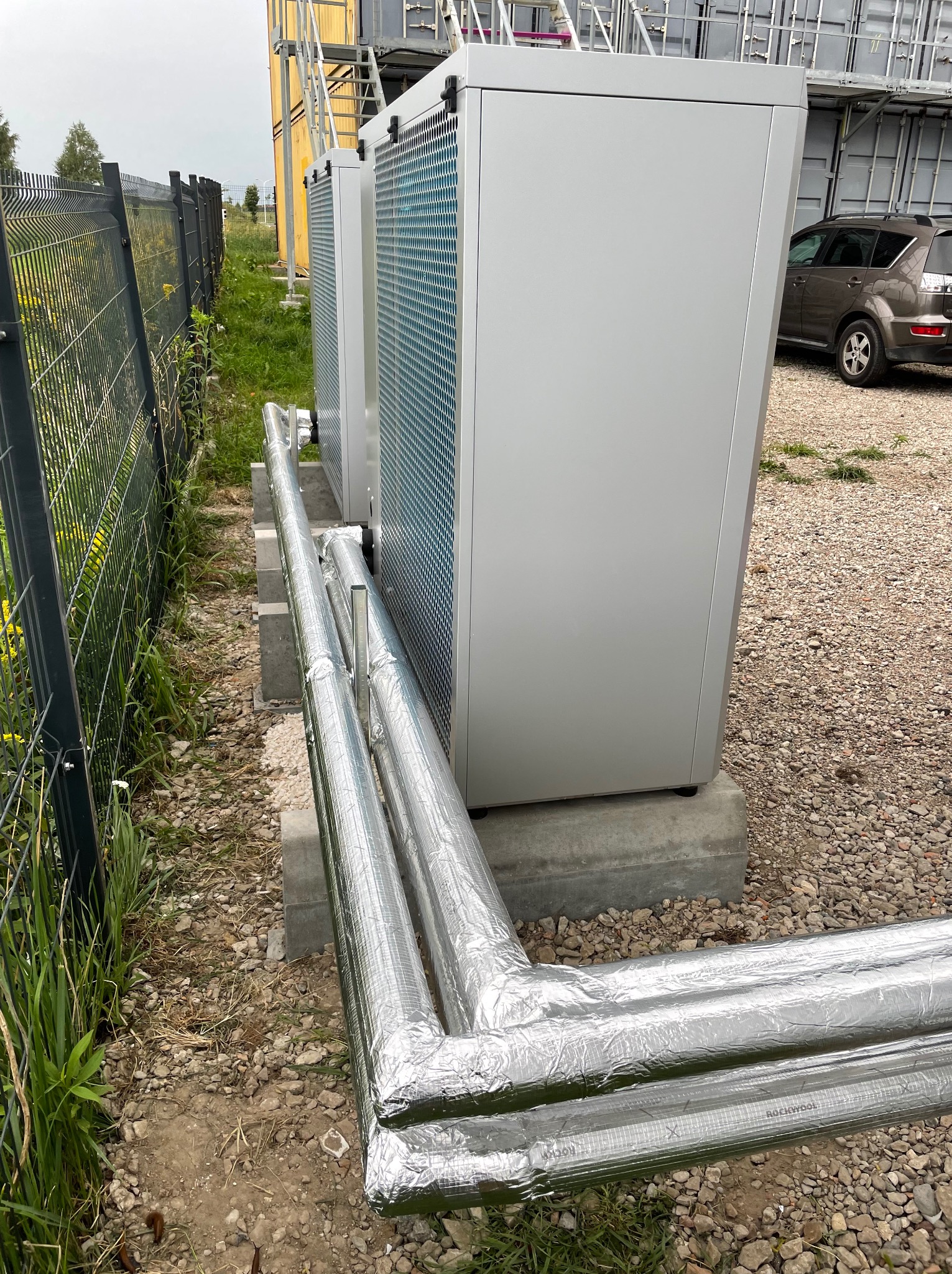 Dwie szare pompy ciepła na betonowych podstawach, z widocznymi rurami izolowanymi folią aluminiową, ułożonymi na żwirowym podłożu obok ogrodzenia z siatki.