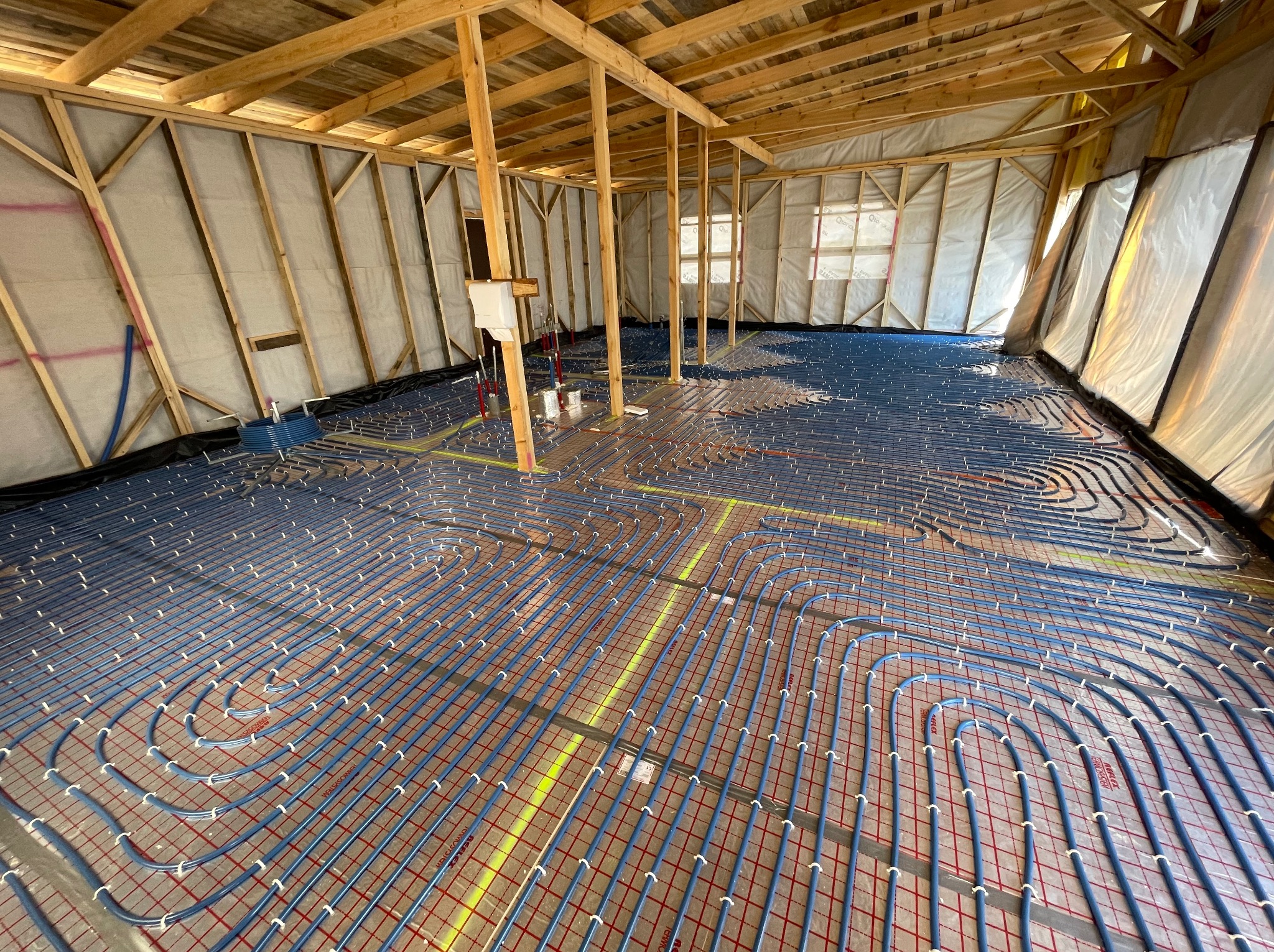 Widok z góry na rozłożone niebieskie rury systemu ogrzewania podłogowego na folii z czerwoną siatką w surowym stanie budowy, widoczne drewniane elementy konstrukcyjne ścian i dachu.