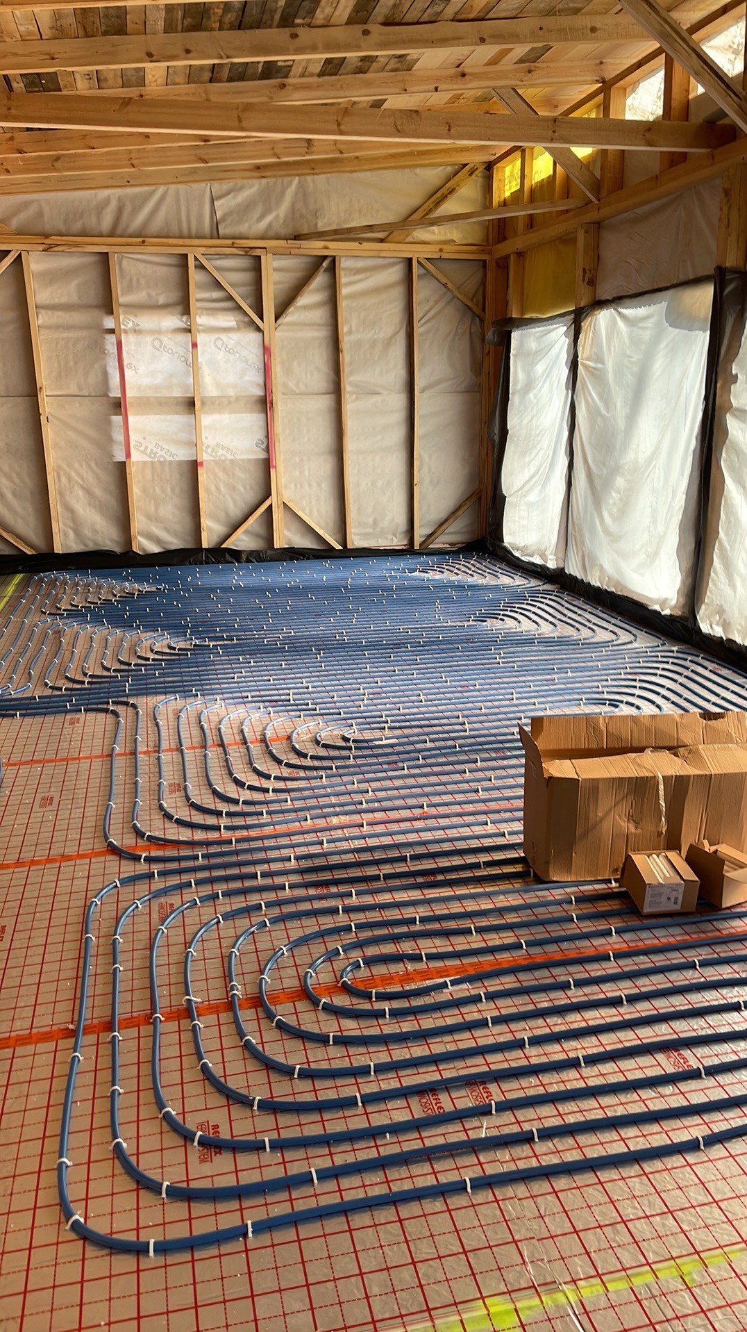 Niebieskie rury ogrzewania podłogowego, mocowane białymi uchwytami, rozłożone na podkładzie z czerwoną kratką w surowym wnętrzu budynku z drewnianą konstrukcją dachu i ścian.