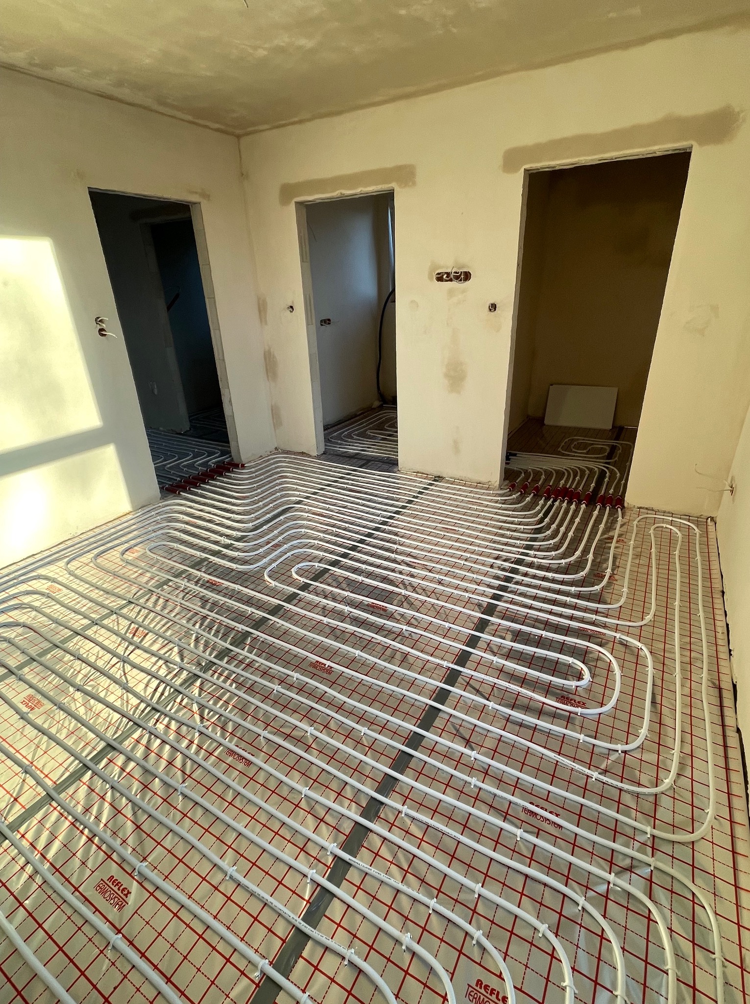 Białe rury ogrzewania podłogowego ułożone na folii refleksyjnej z czerwoną siatką, w surowym wnętrzu z trzema otworami drzwiowymi i naturalnym oświetleniem słonecznym.