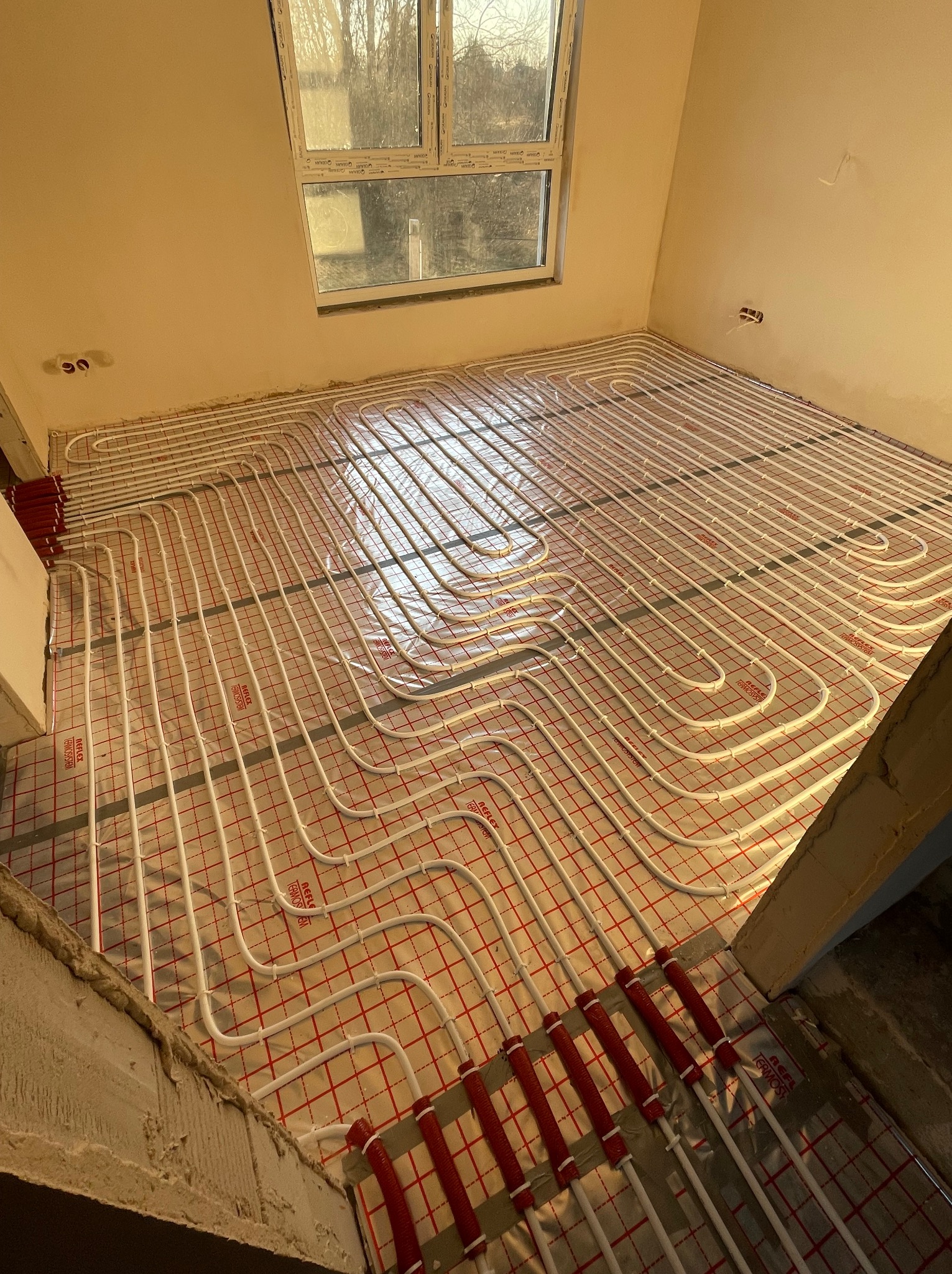 Montaż ogrzewania podłogowego: białe rurki ułożone na czerwonej siatce z folią refleksyjną w surowym wnętrzu, widoczne okno i fragment ściany.
