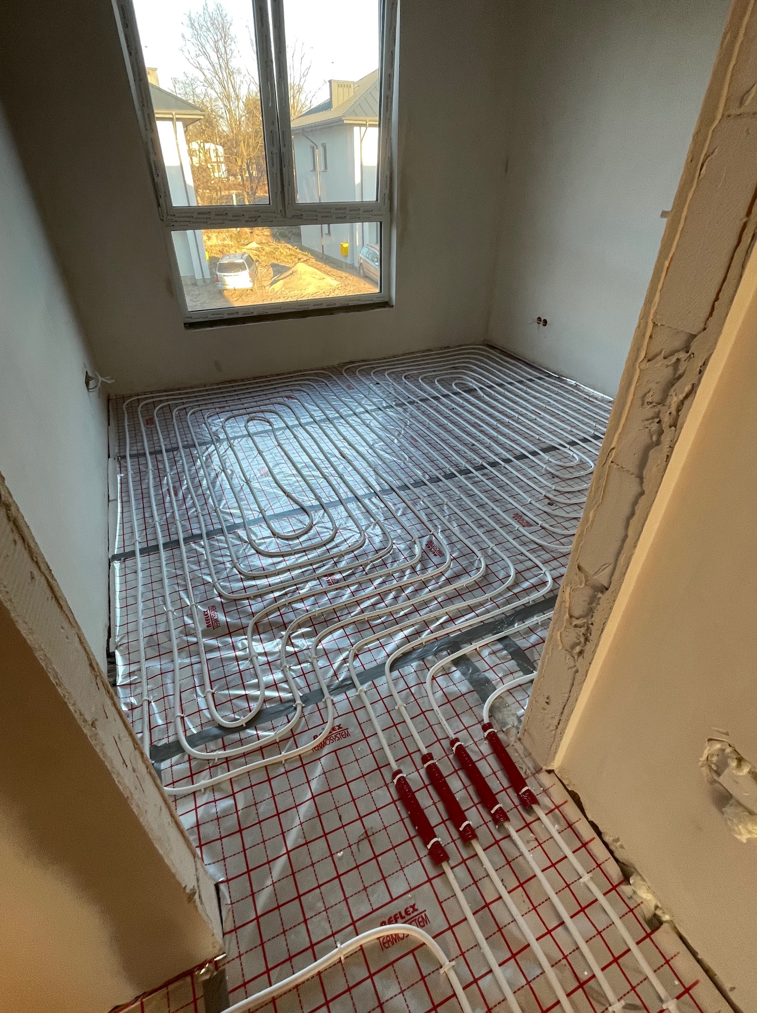 Instalacja ogrzewania podłogowego: białe rurki ułożone na folii refleksyjnej z czerwoną siatką w surowym wnętrzu z widokiem na dom z ogrodem przez okno.