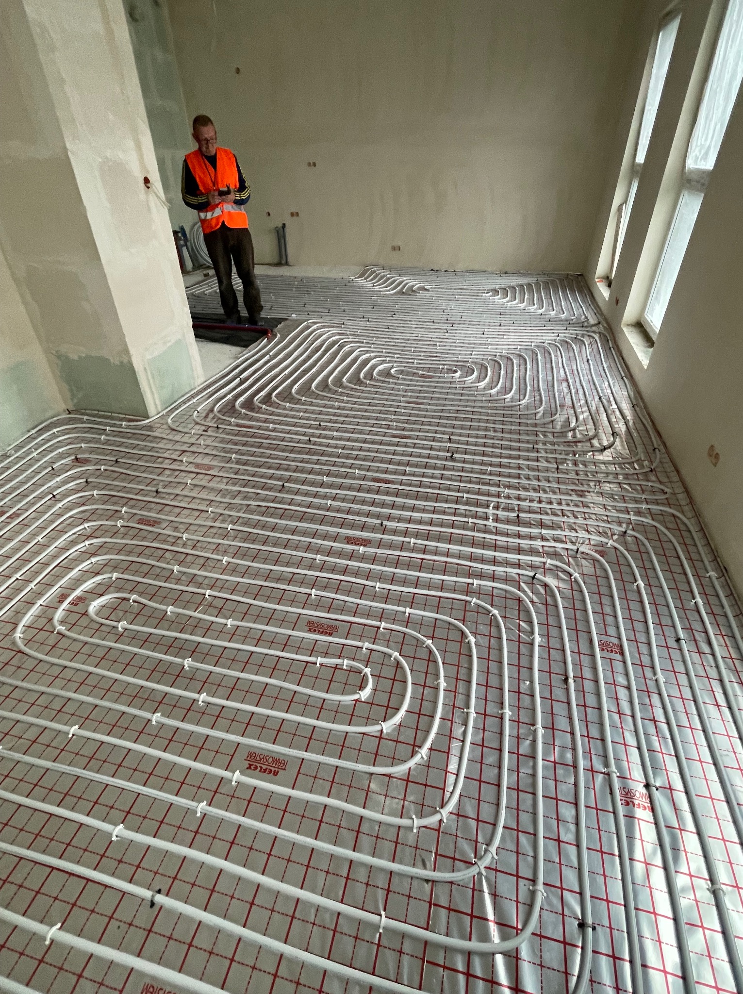 Układanie rur ogrzewania podłogowego na folii z czerwoną kratką, widoczny instalator w pomarańczowej kamizelce z telefonem w surowym wnętrzu.