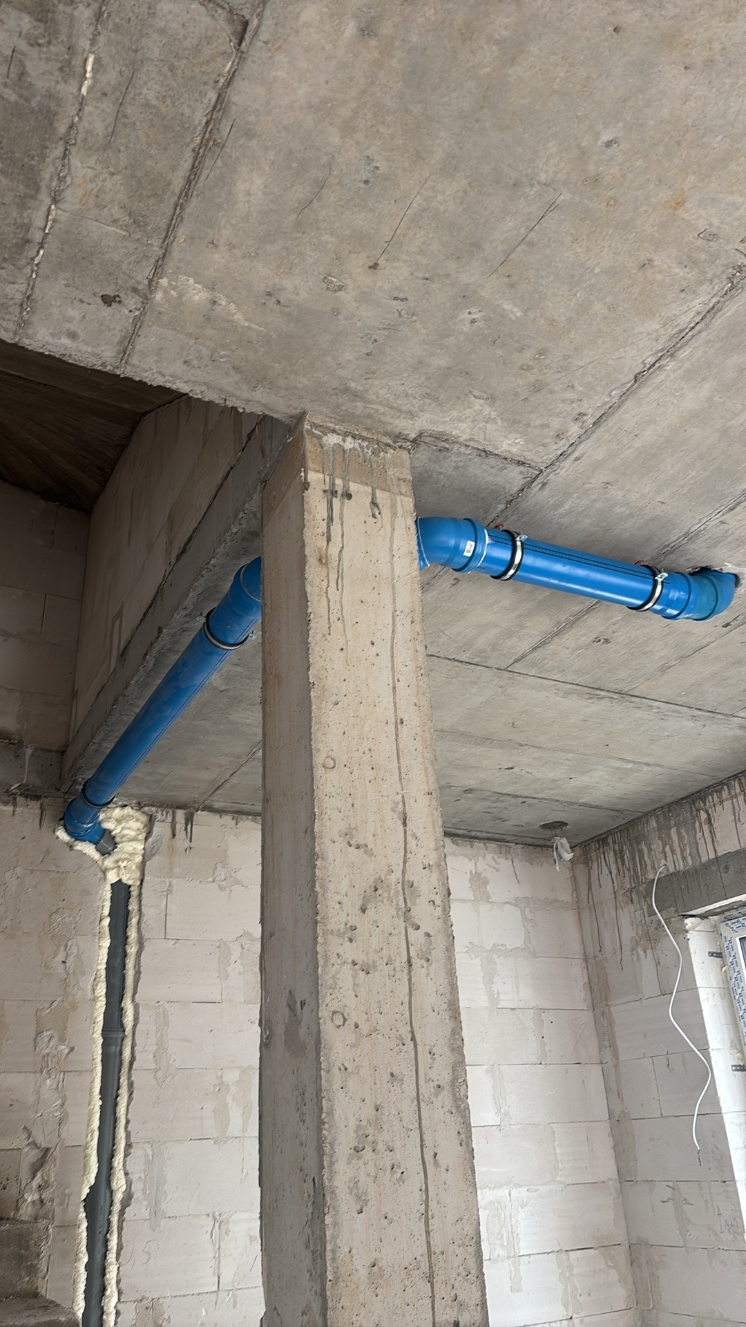 Niebieskie rury instalacji wodnej zamocowane do surowego betonowego stropu w surowym stanie deweloperskim budynku.