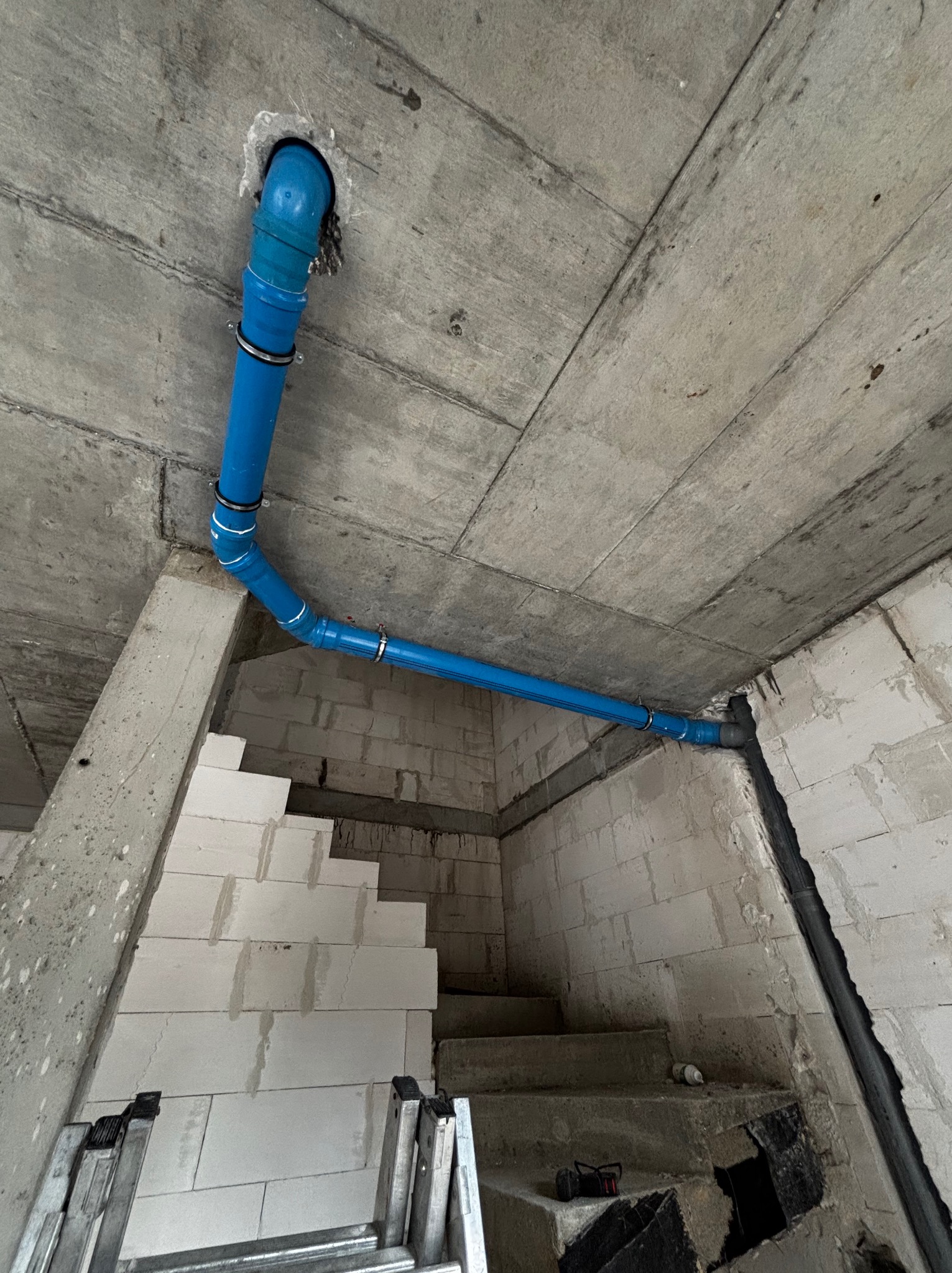 Niebieska rura instalacji wodnej przymocowana do surowego betonowego sufitu w surowym stanie, widok z dołu na schody i drabinę.
