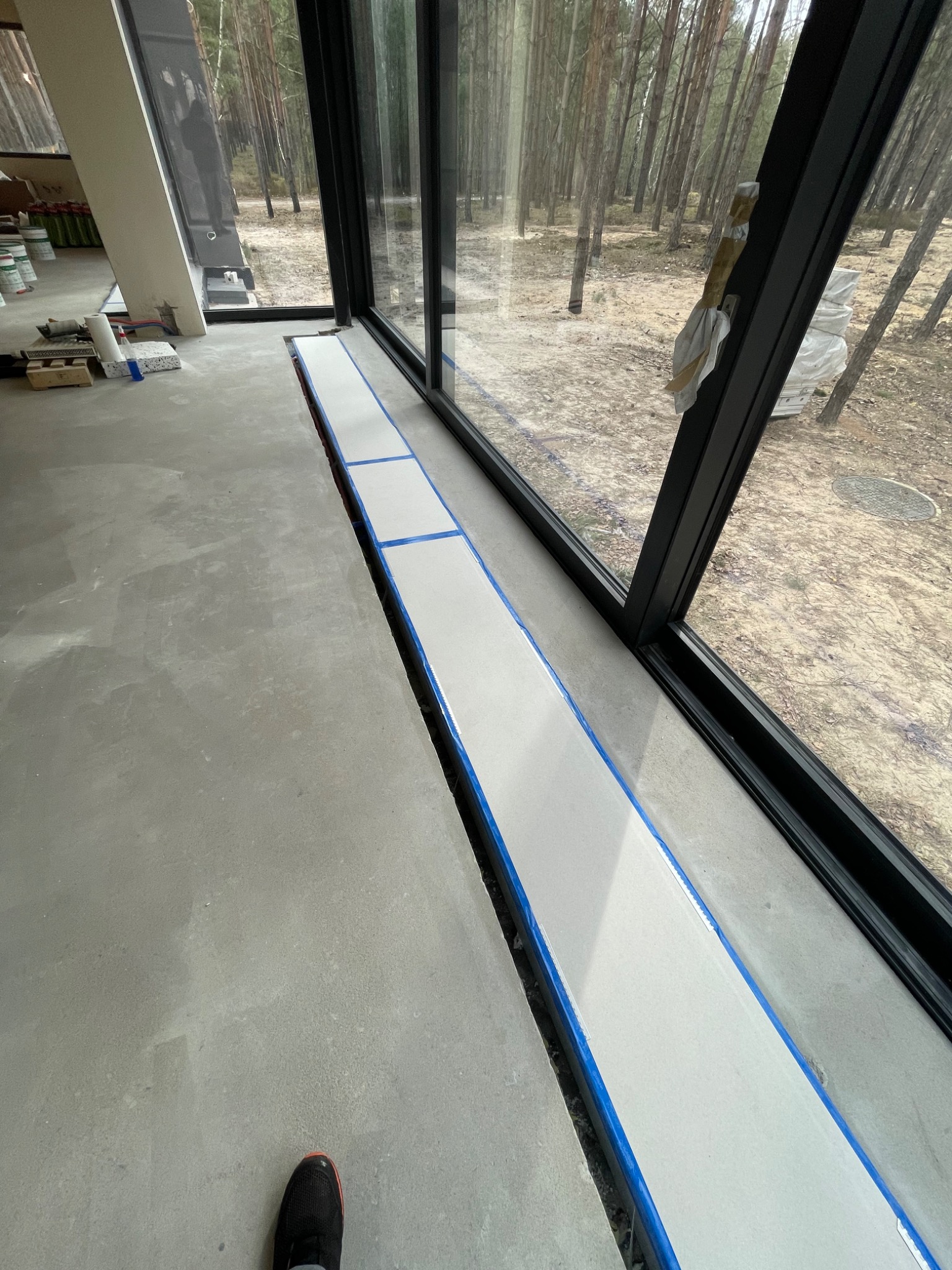 Przygotowanie do montażu ogrzewania podłogowego w nowoczesnym wnętrzu z widokiem na las; widoczna wylewka z taśmą zabezpieczającą przy panoramicznym oknie.