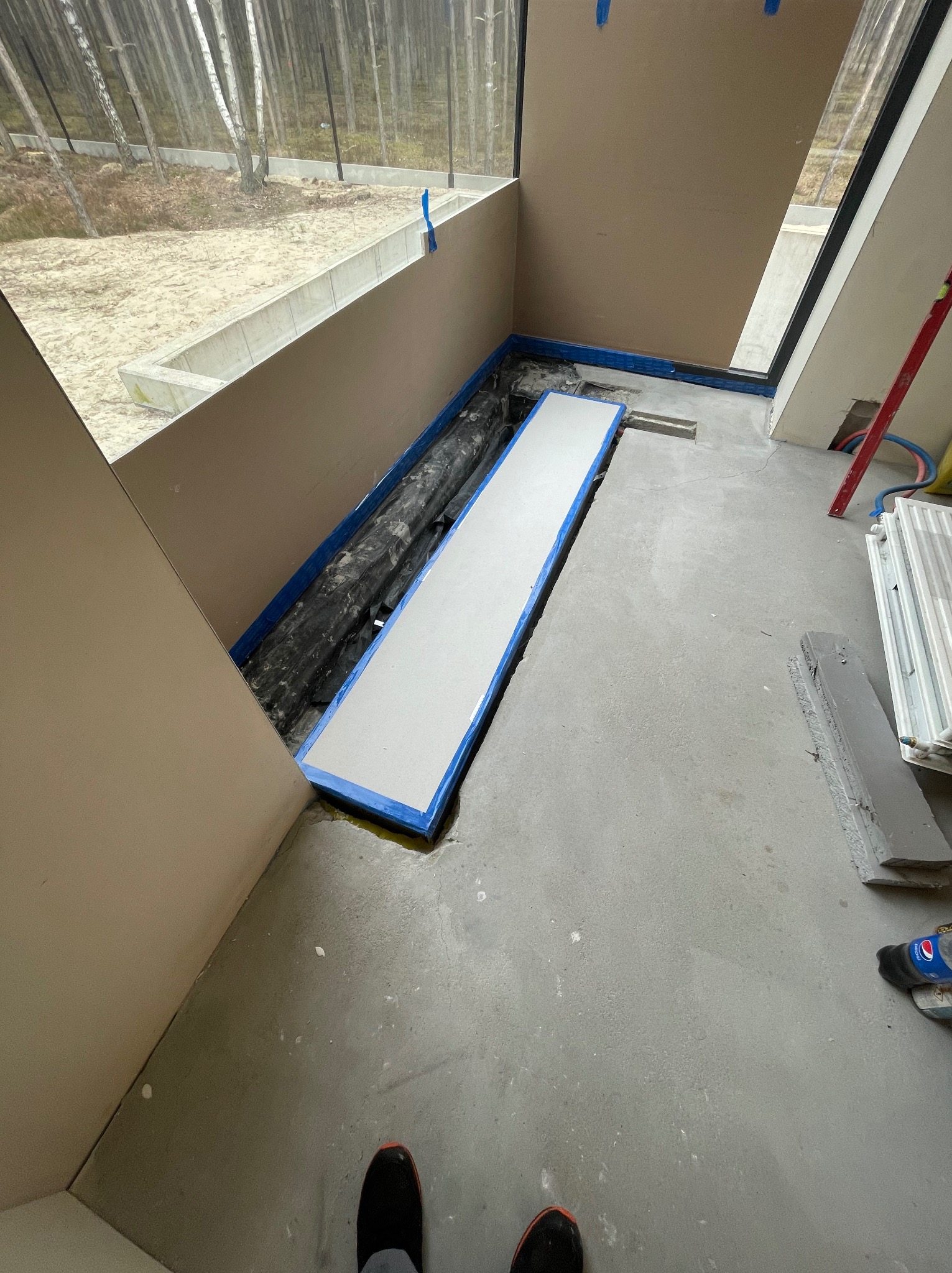 Przygotowanie podłoża pod ogrzewanie podłogowe w nowoczesnym wnętrzu z dużym oknem, widoczne rury w izolacji, płyta styropianowa z niebieską taśmą, poziomica, grzejnik i butelka Pepsi.