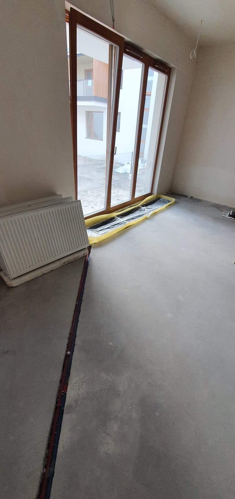 Przygotowanie do montażu ogrzewania podłogowego: widoczne rury w wylewce, folia izolacyjna pod oknem i grzejniki czekające na instalację w surowym wnętrzu.