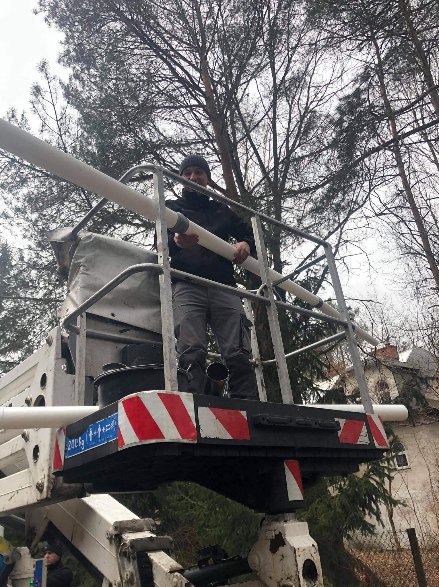 Pracownik na podnośniku koszowym w Warszawie instaluje rury, prawdopodobnie wentylacyjne, na tle drzew i budynku.