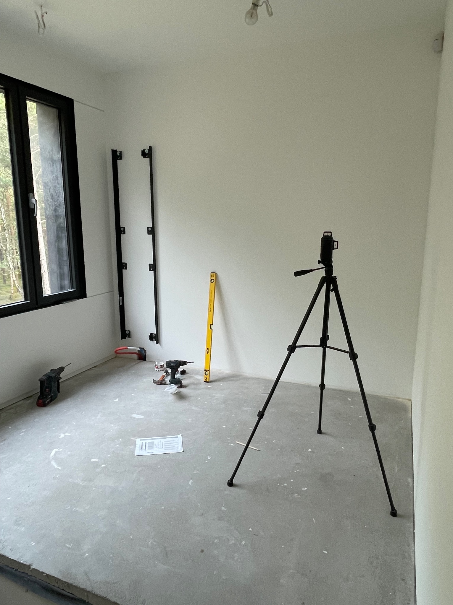 Wnętrze w trakcie montażu instalacji: laser krzyżowy na statywie, poziomica, wiertarki i listwy montażowe na ścianie, surowy beton na podłodze.